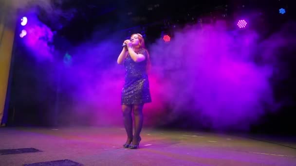 Una joven cantante pelirroja con un micrófono en las manos con un vestido negro canta en el escenario en pleno crecimiento — Vídeo de stock