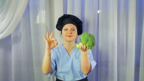Seorang wanita memasak di celemek tersenyum, memegang brokoli di tangannya dan menunjukkan tangannya OK — Stok Video