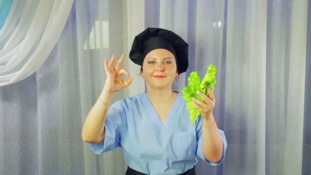 Mujer cocinera en delantal sonríe, sostiene ensalada verde en la mano y muestra la mano OK . — Vídeo de stock