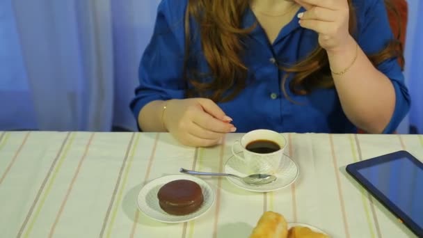 Жінка в кафе за столом п'є каву з десертом і читає новини на планшеті — стокове відео