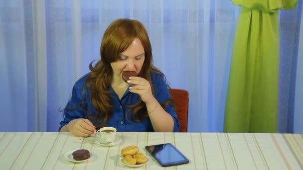 Jovem mulher de cabelos castanhos em um café em uma mesa bebendo café com sobremesa e lendo as notícias no tablet — Vídeo de Stock