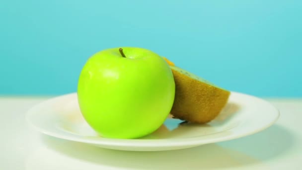Целые зеленые яблоко и половина киви и апельсин на белой тарелке вращаются в круге — стоковое видео