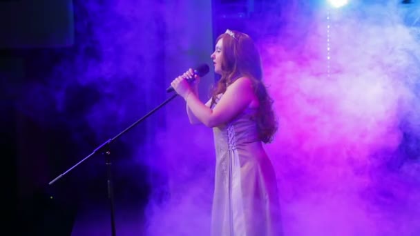 Μια νεαρά κοκκινομάλλα τραγουδίστρια τραγουδά στη σκηνή με επαγγελματικό φωτισμό και ψεκασμούς καπνού. Πλευρά σκοποβολής — Αρχείο Βίντεο