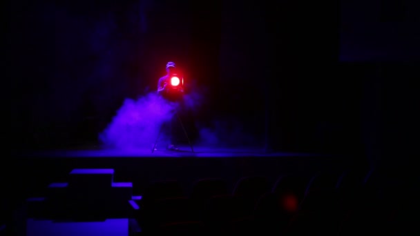 Un illuminatore uomo dirige un fascio di luce da un riflettore di profilo dal palco a una palestra in nuvole di fumo — Video Stock