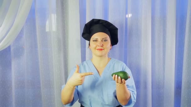 Uma cozinheira sorri, segura um abacate na mão e aponta para ele . — Vídeo de Stock