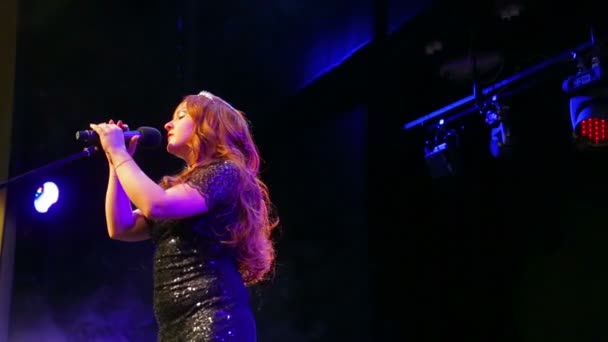 Una giovane attrice dai capelli rossi in un abito nero lucido canta sul palco al microfono in un soffio di fumo — Video Stock