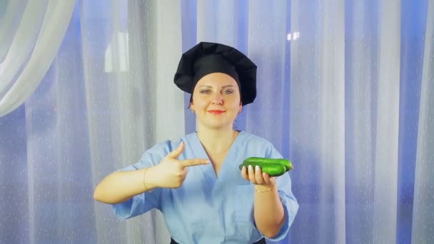Женщина-повар в фартуке улыбается, держит в руке огурцы и указывает на него — стоковое видео