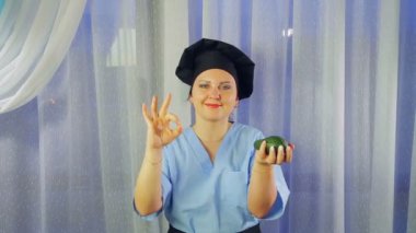 Önlüklü kadın gülümseyerek yemek pişirir, elinde avokado tutar ve elleriyle OK 'u gösterir.