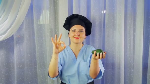 Mujer cocinera en delantal sonríe, sostiene aguacate en su mano y muestra OK con su mano — Vídeo de stock