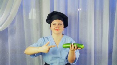 Önlüklü bir kadın yemek yapıyor, elinde kabak tutuyor ve ona işaret ediyor.