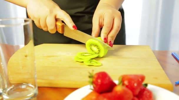 Una mujer está cortando un kiwi maduro con un cuchillo en una tabla de madera con un cuchillo — Vídeo de stock