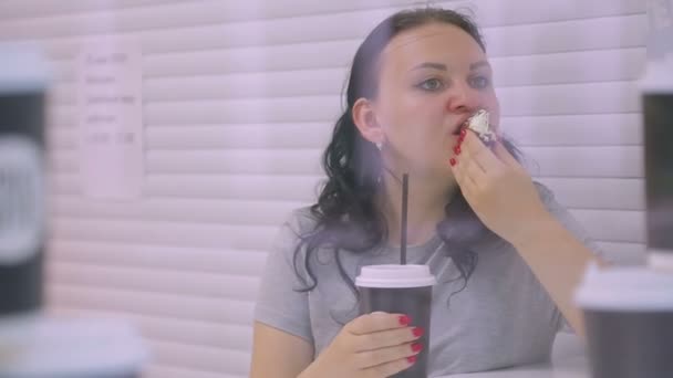 Een brunette vrouw in een café drinkt koffie van een donker glas en eet een cake. Schieten door het glas — Stockvideo