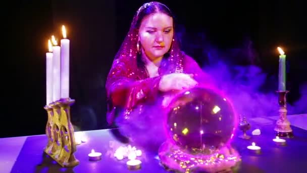 Una mujer gitana en un salón mágico en bocanadas de humo adivina en una bola de cristal y un signo de bitcoin aparece en ella . — Vídeo de stock