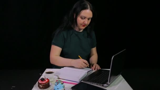 Une femme dans un café boit le café d'une tasse blanche sur un fond noir et écrit dans un journal tout en regardant dans un ordinateur — Video