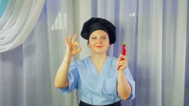 Woman cook in apron smiles, holds chilli pepper in her hand and shows her hand OK
