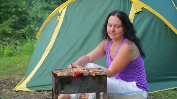 Молодая женщина в футболке сидит в лесу на зеленом фоне палатки, готовит ужин в своей жаровне — стоковое видео