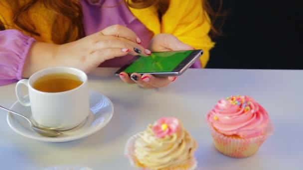 A menina em um café na mesa com bolos folheando as notícias no smartphone . — Vídeo de Stock