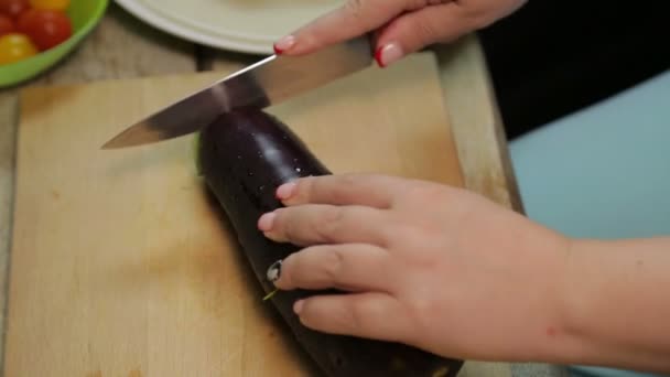 La mano femenina corta una berenjena mediana con un cuchillo en una tabla de madera — Vídeo de stock