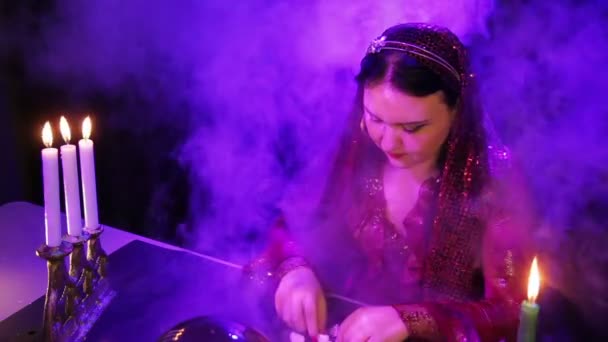 Циганка в червоній сукні в чарівному салоні в диму при свічках читає майбутнє на каменях — стокове відео