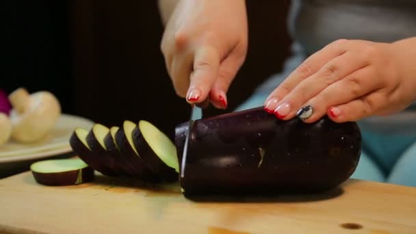 Weibliche Hand schneidet eine mittlere Aubergine mit einem Messer auf einem Holzbrett — Stockvideo