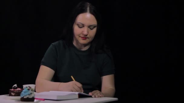 Eine Frau in einem Café trinkt Kaffee aus einer weißen Tasse auf schwarzem Hintergrund und schreibt in ein Tagebuch — Stockvideo