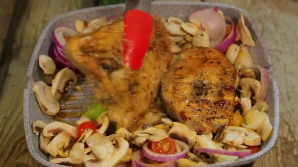 Mão feminina vira bifes de peixe grelhados com pinças e mistura cogumelos com cebola . — Vídeo de Stock