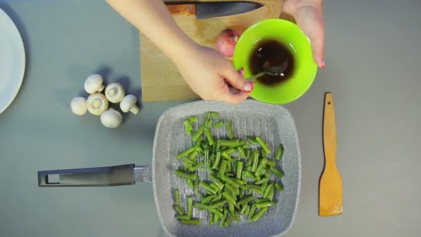 Een vrouw is het roeren van een sojasaus met een lepel en het gieten van groene bonen op een pan — Stockvideo