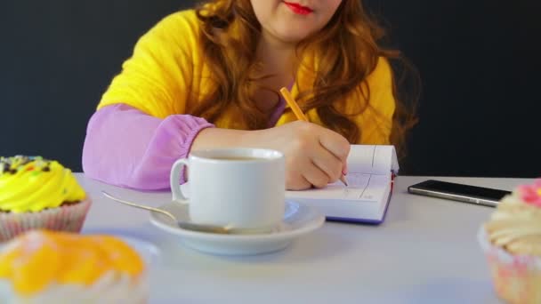 A lány a kávézóban az asztalnál írja a naplóban — Stock videók