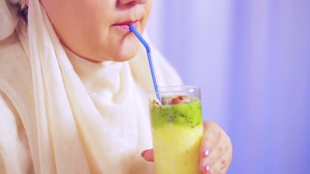 Una joven musulmana con una bufanda ligera sostiene un vaso con un batido de frutas en las manos y bebe de una pajita — Vídeos de Stock