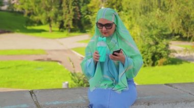 Parkta hafif bir eşarp ve güneş gözlüğü genç bir Müslüman kadın kahve içiyor ve messenge içinde