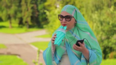 Parkta hafif bir eşarp genç bir Müslüman kadın kahve içiyor ve kulaklık ile haberci konuşuyor