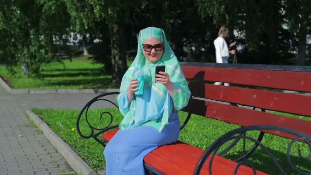 Μια νεαρή μουσουλμανική γυναίκα με ένα ελαφρύ κασκόλ χύση σε ένα παγκάκι στο πάρκο και πίνει καφέ και μιλά στον αγγελιοφόρο. — Αρχείο Βίντεο