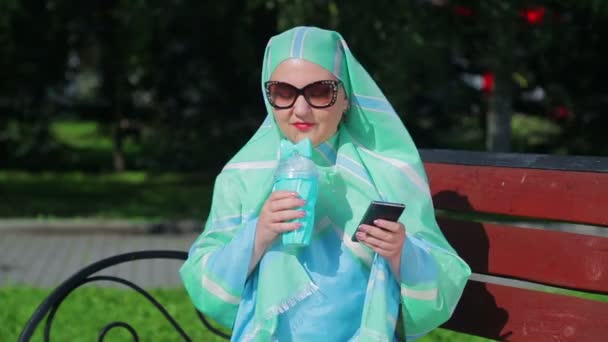Een jonge moslim vrouw in een lichte sjaal en zonnebril zit op een bankje in het Park en drinkt koffie en praat in de boodschapper — Stockvideo