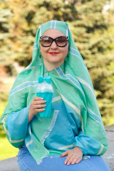 Eine junge muslimische Frau mit hellem Schal und Brille im Park trinkt Kaffee. — Stockfoto