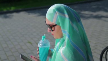 Hafif bir eşarp genç bir Müslüman kadın parkta bir bankta oturur ve kahve ve haberci sohbet. Yan görünüm