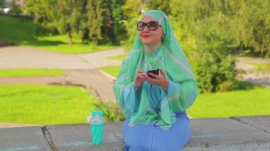Parkta hafif bir eşarp ve güneş gözlüğü genç bir Müslüman kadın kahve içiyor ve haberci