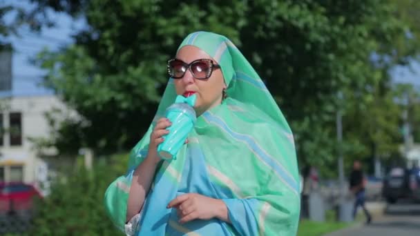Parkta hafif bir atkı ve güneş gözlüğü takan genç bir Müslüman kadın kahve içiyor.. — Stok video