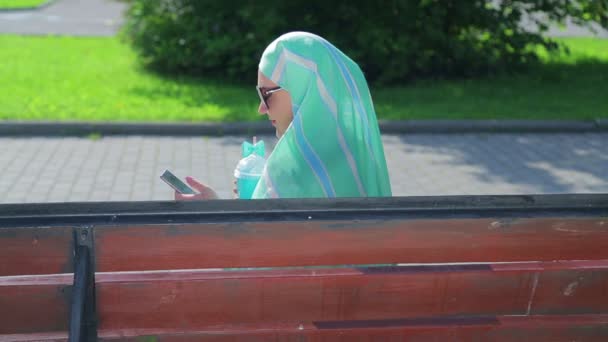 A young Muslim woman in a light scarf sits on a bench in the park and drinks coffee and talks in the messenger. Side view — Stock Video