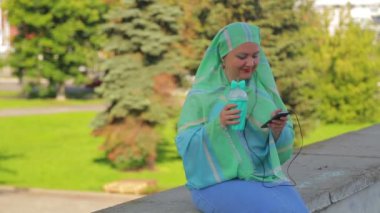 Parkta hafif bir eşarp genç neşeli Müslüman kadın kahve içiyor ve kulaklık haberci konuşuyor