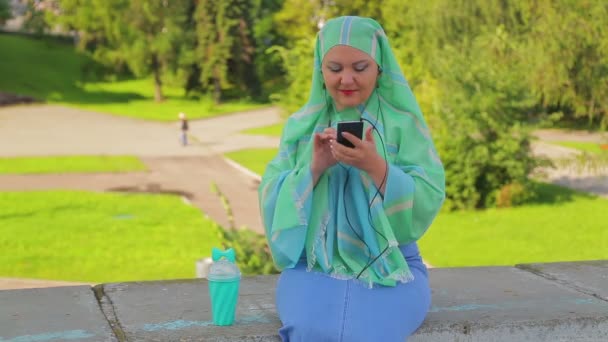 Een jonge moslim vrouw in een groene sjaal in het Park drinkt koffie en is in de Messenge — Stockvideo
