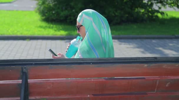 Una giovane donna musulmana con una sciarpa leggera siede su una panchina nel parco e beve caffè e parla nel messaggero. Vista laterale — Video Stock