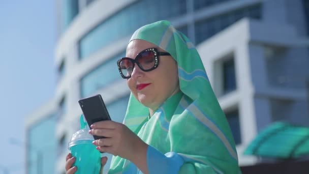 Una giovane donna musulmana in una sciarpa leggera e occhiali da sole sullo sfondo di un edificio moderno — Video Stock