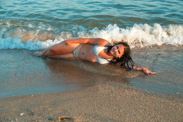 Uma jovem mulher está deitada na areia junto ao mar em um maiô branco nas ondas costeiras. . — Fotografia de Stock