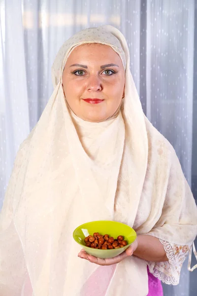 Eine junge muslimische Frau in einem hellen Schal steht am Tisch mit Früchten und hält einen Teller mit Nüssen in der Hand. — Stockfoto