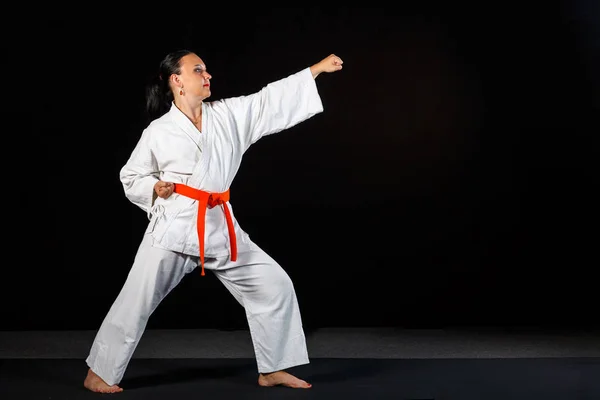A young brunette woman in a white kimono doing karate on a black background in full growth. — Stock Photo, Image