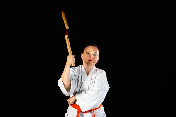 En ung brunett kvinna i en vit kimono är engagerad med nunchucks på en svart bakgrund. — Stockfoto