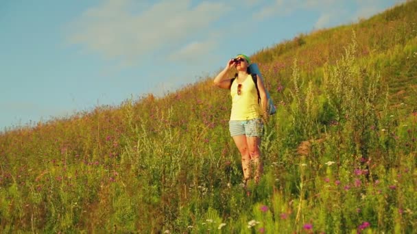Mujer excursionista en una colina con una mochila detrás de ella, mirando a través de los prismáticos — Vídeos de Stock