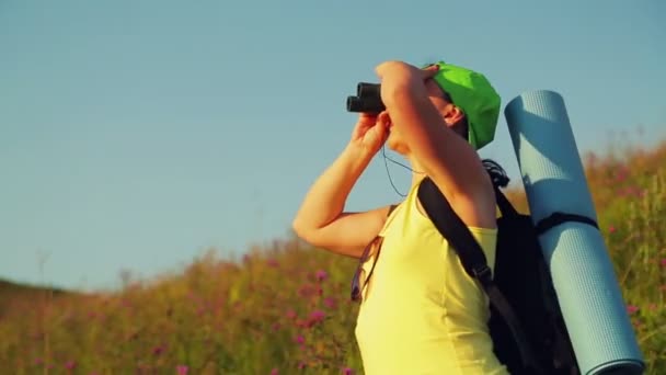 Mujer excursionista en una colina con una mochila detrás de ella, mirando a través de prismáticos en los lugares de interés . — Vídeos de Stock