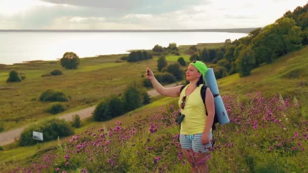 Kvinna turist på en kulle med en ryggsäck bakom honom och kikare gör ett foto på telefonen. — Stockvideo