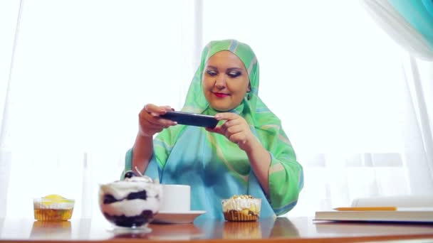 Een jonge moslim vrouw in een groene sjaal in een café Foto's gebak op de telefoon — Stockvideo